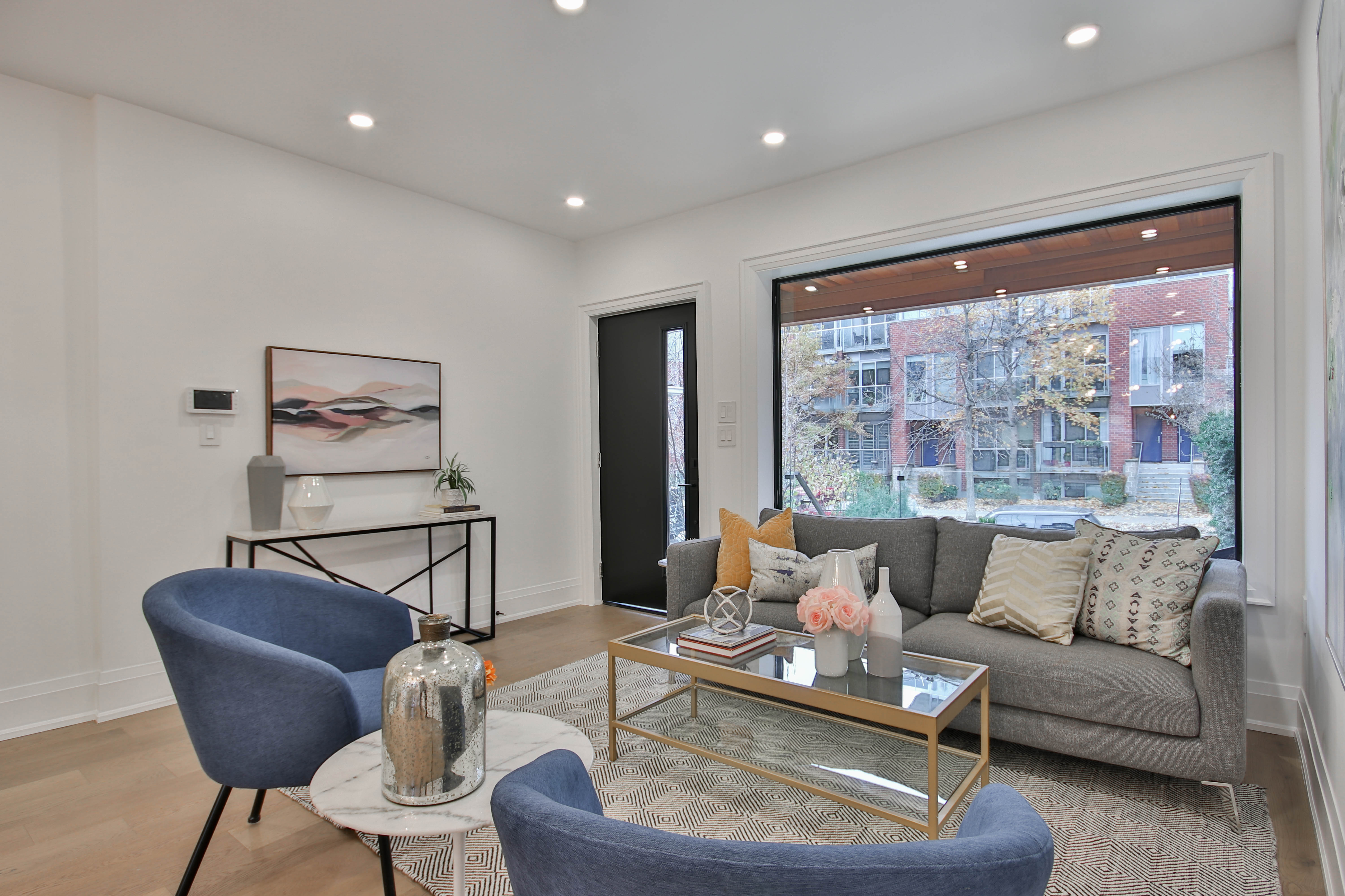 An elegant residential interior designed by a Toronto architecture studio, featuring an open‑concept living and dining area with floor‑to‑ceiling windows, pale oak floors, and a sculptural black steel staircase with glass guardrails. A minimalist, built‑in wall unit in matte white conceals storage, while a low stone-clad fireplace anchors the space. Soft, diffused daylight filters through sheer curtains, creating refined highlights on the textured upholstery and subtle shadows along the ceiling. Shot at eye level with sharp focus throughout, the composition looks from the living area toward a distant kitchen, conveying flow and spatial clarity. The atmosphere is calm, luxurious, and meticulously detailed, in a clean photographic style.